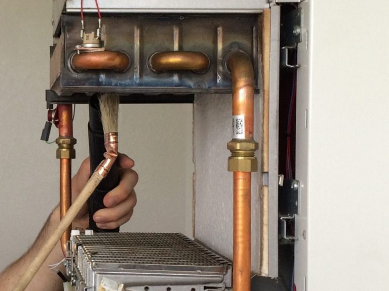 Main en train de manipuler un pinceau et un tuyau d'aspirateur pour nettoyer une chaudière à gaz